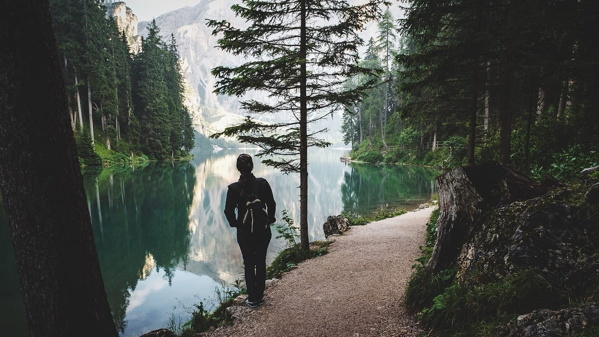 A person stands on a gravel path beside a still mountain lake surrounded by forest