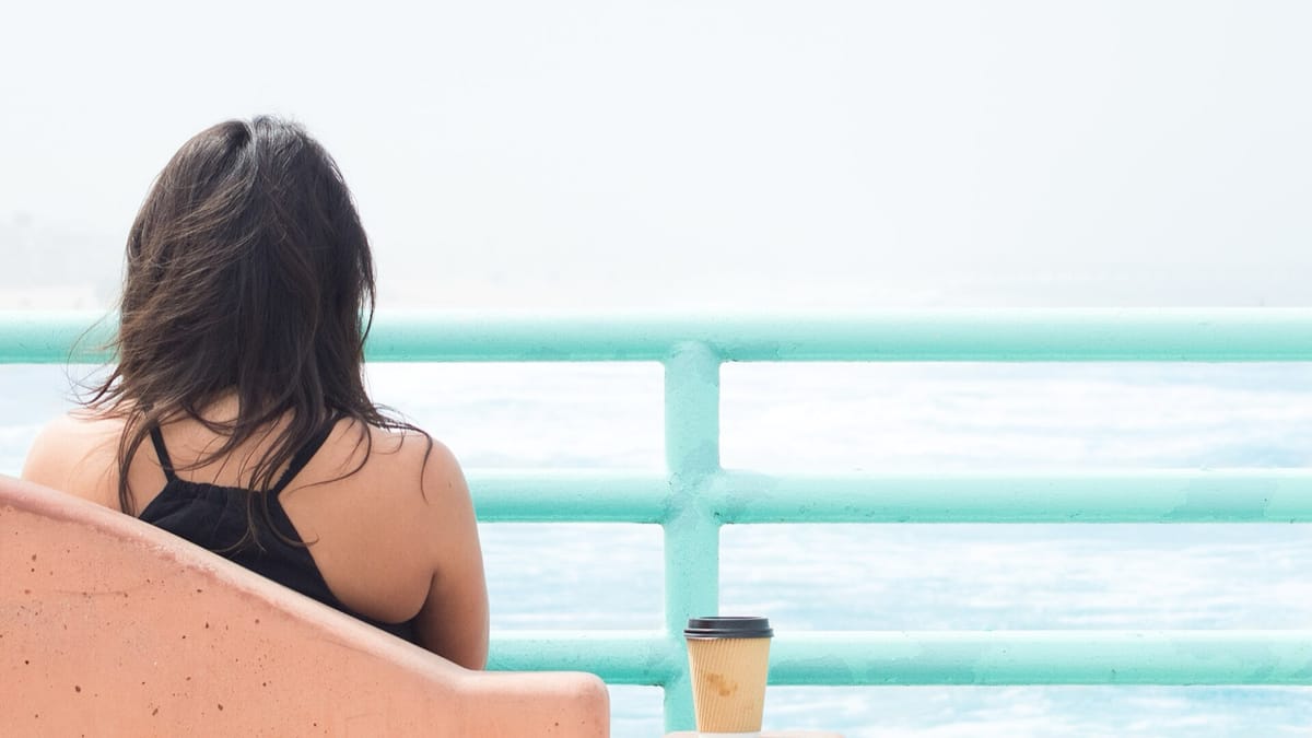 A woman sits quietly facing the ocean from a seaside railing, a coffee cup beside her
