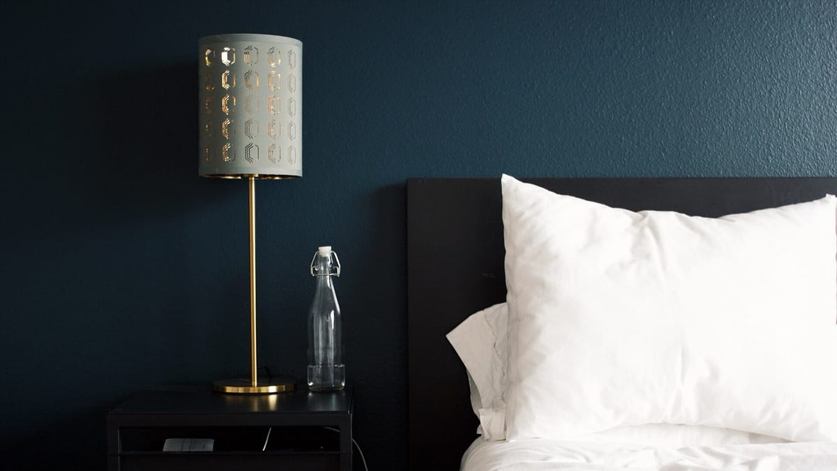 A bedside lamp and a glass water bottle beside a bed with white pillows against a dark teal wall